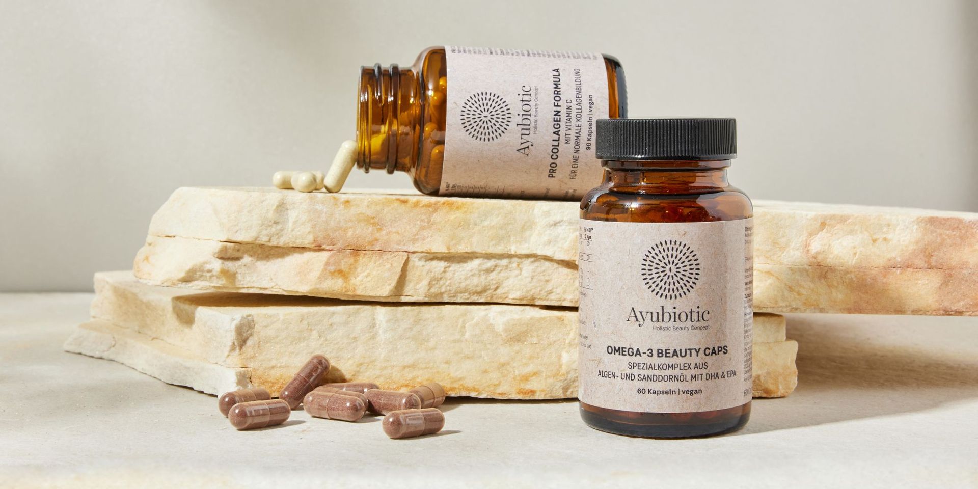 Braune Ayubiotic-Nahrungsergänzungsflaschen und Kapseln auf hellem Steinuntergrund.