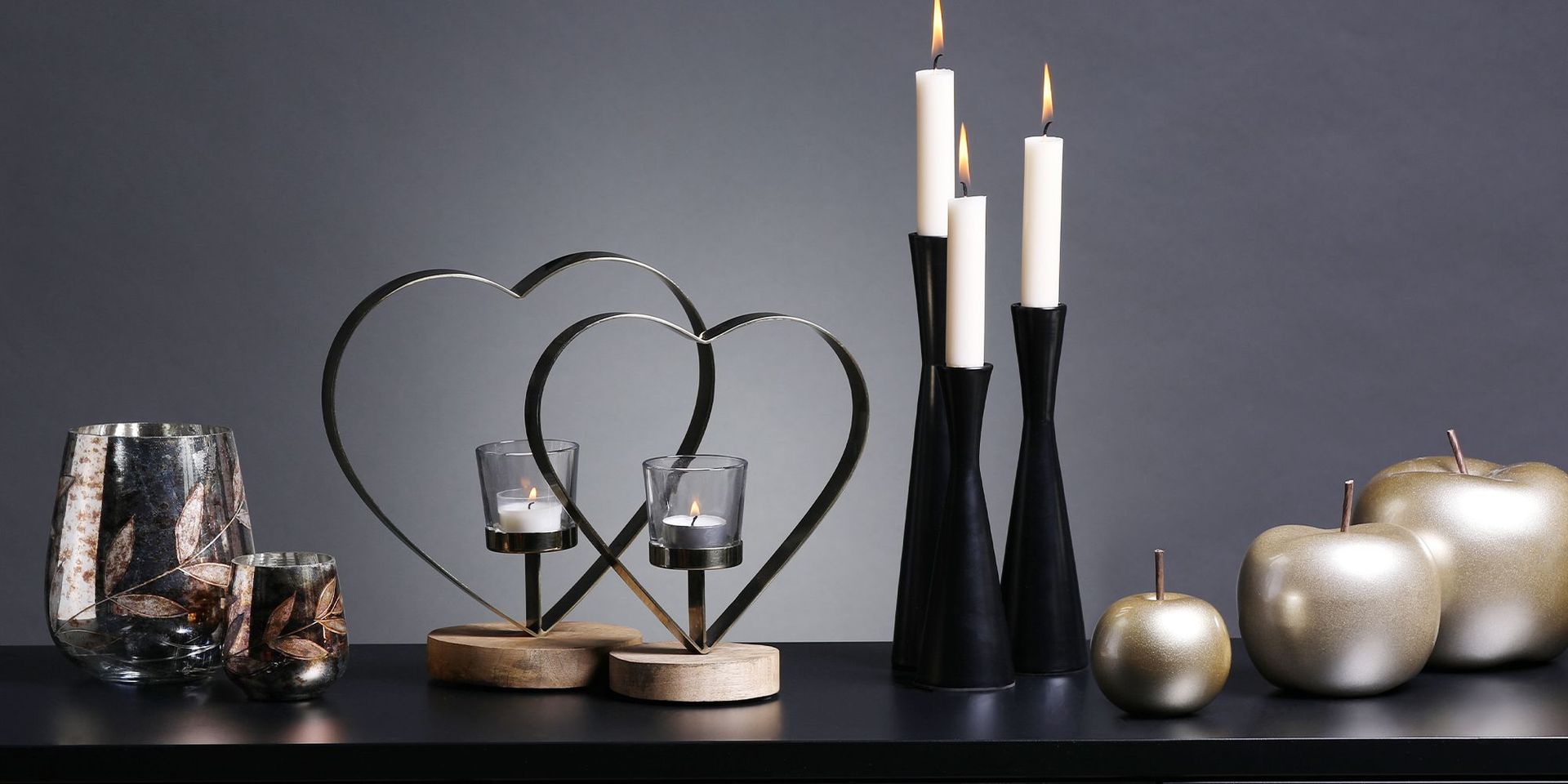 Moderne Kerzenhalter in Herzform und schlanke schwarze Kerzenständer mit brennenden Kerzen auf schwarzem Tisch.